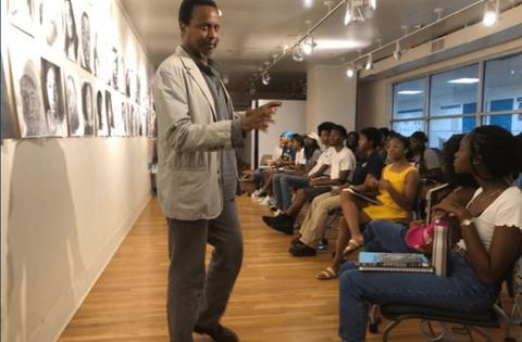 Black male professor wearing white blazer speaking in front of a group of students